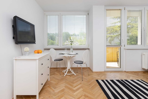 A small, bright room with a white desk, two chairs, a television, and large windows.