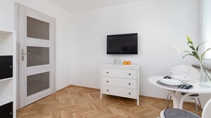 Une pièce dotée d’un parquet en bois à motifs chevrons, d’une commode blanche avec des tiroirs, d’un téléviseur à écran plat fixé au-dessus, d’une table à manger avec des chaises et d’une fenêtre avec des stores.