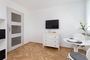 A room with a herringbone wooden floor, a white dresser with drawers, a flat-screen TV mounted above it, a dining table with chairs, and a window with blinds.