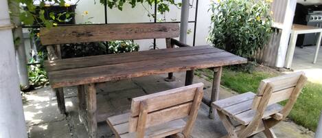 Una mesa de madera al aire libre con bancos, rodeada de follaje y un jardín.