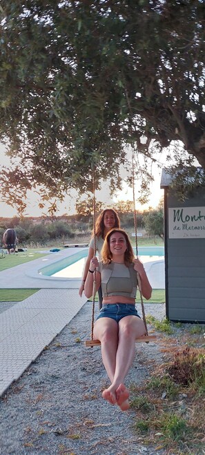 Zwei Personen sitzen auf einer Schaukel unter einem Baum, im Hintergrund ein Schild mit der Aufschrift 'Mont de la Mer'.