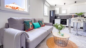 A modern living room with a grey sofa, colorful cushions, a round coffee table, and a view of the kitchen in the background.