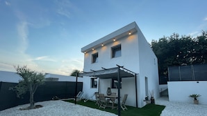 A modern two-story house with a patio area featuring a table and chairs, a pergola, and a small tree.