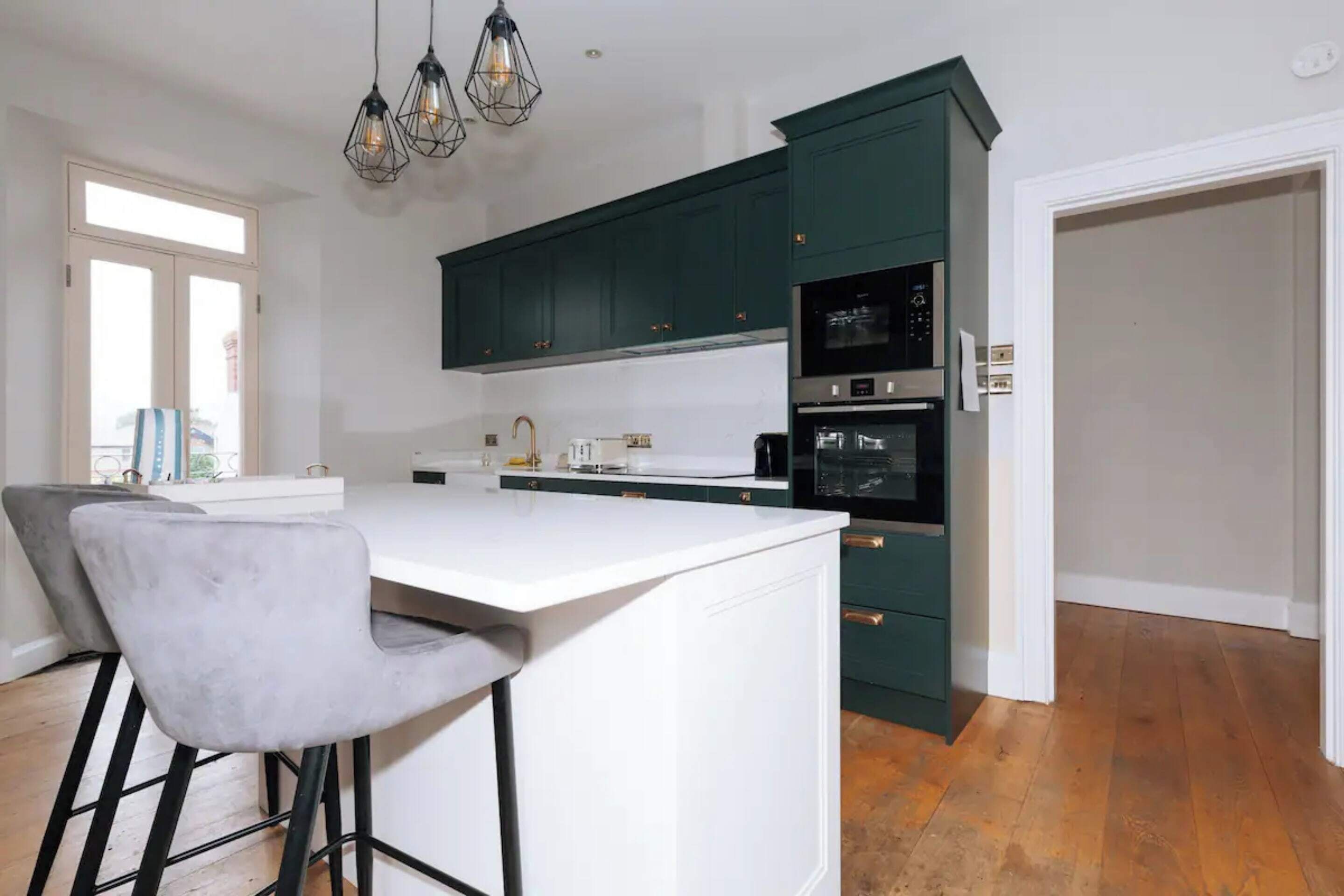 A modern kitchen with dark green cabinets, a white countertop, and a bar stool.