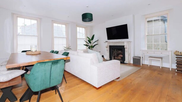 A spacious living room with a wooden floor, a white sofa, a dining table with chairs, a fireplace, and a television mounted on the wall. - Chic & Characteristic 2BD House - Irishtown (Dublin)