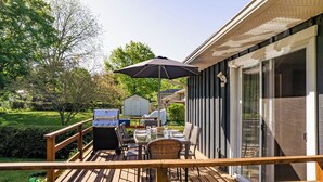 Eine Terrasse mit Grill, Esstisch und Sonnenschirm, mit Blick auf einen Garten mit einem Haus und Bäumen.