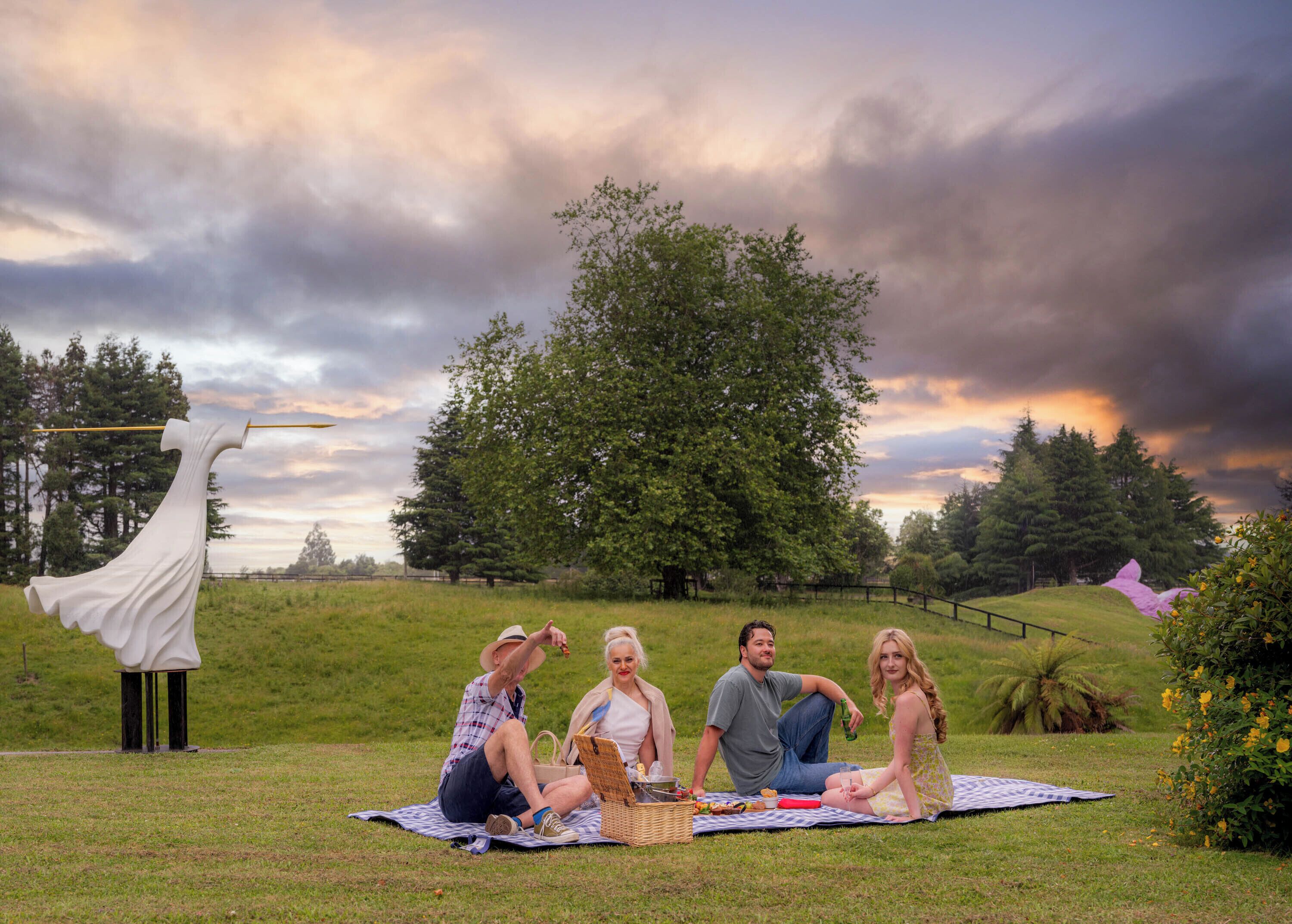 Un grupo de personas en una manta de picnic en un parque con césped, árboles y una escultura.