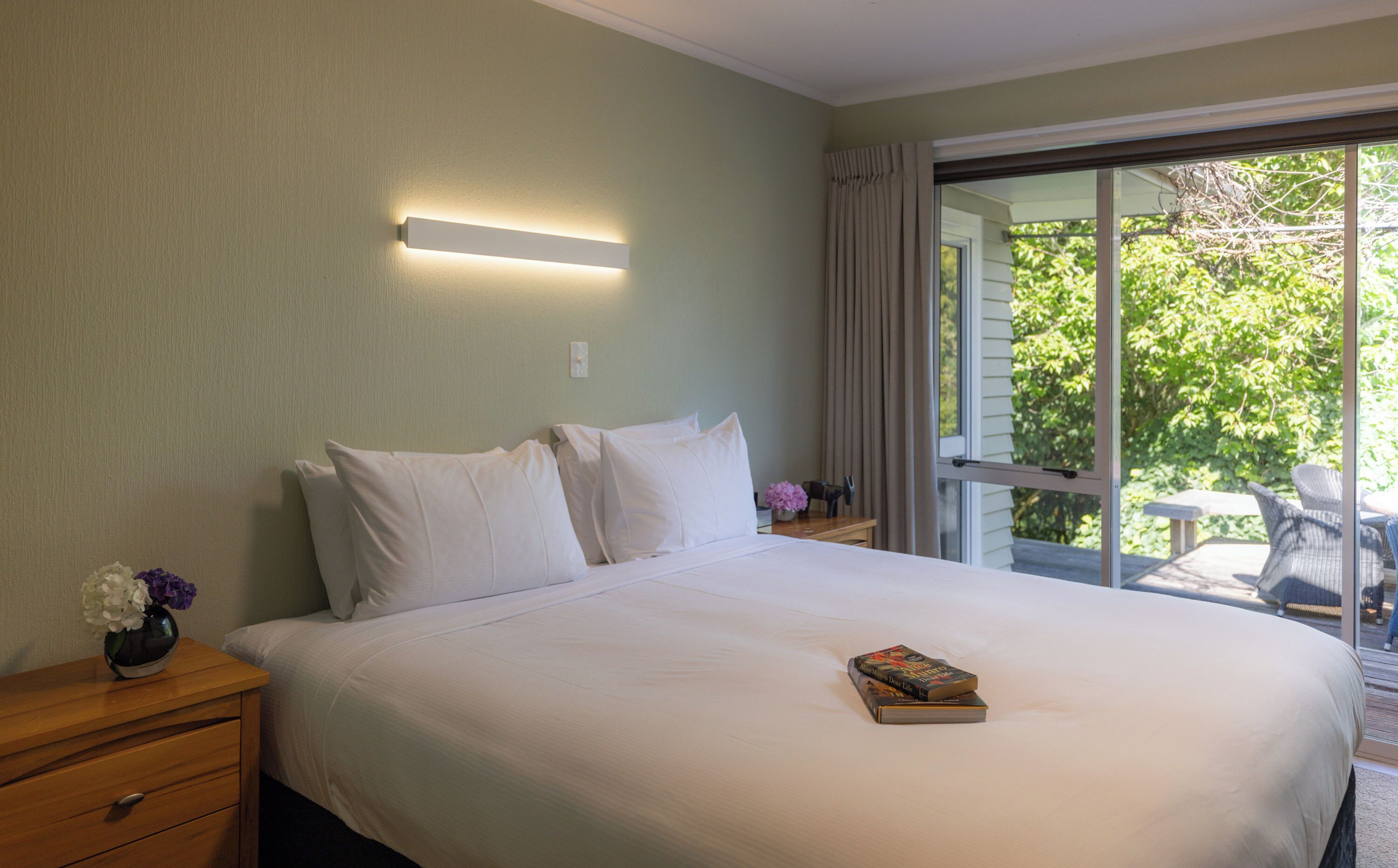 A neatly made bed with white linens and pillows, a bedside table with a vase of flowers, and a book on the bed.