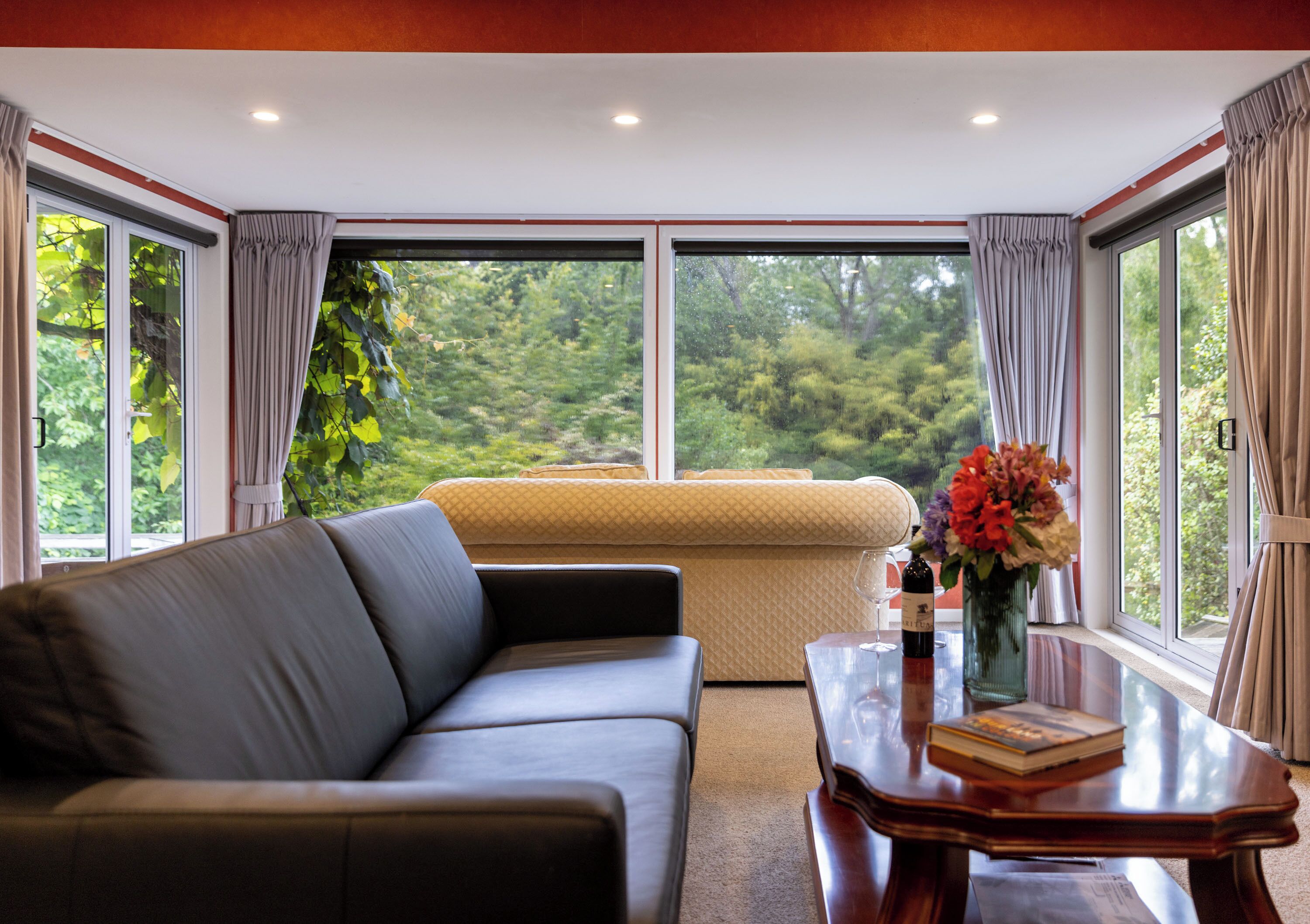 A modern room with a leather sofa, a glass coffee table with a book and wine bottle, and a view of lush greenery through large windows.