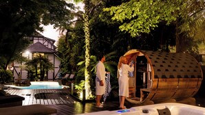 Un área de hidromasaje con una sauna de barril de madera, una piscina y una casa al fondo.