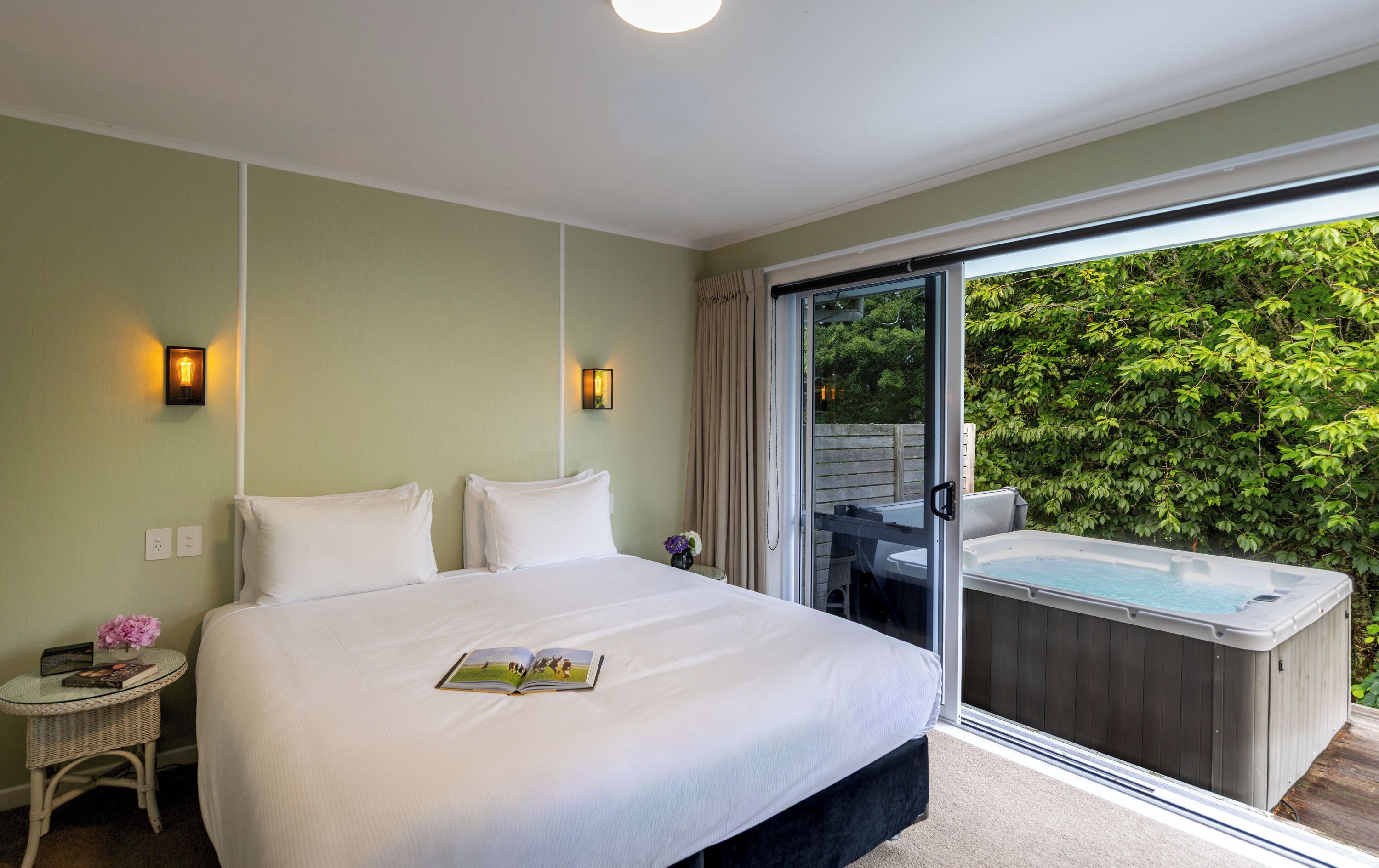 A bedroom with a large bed, two bedside lamps, a small side table with a book and flowers, and a sliding glass door leading to a hot tub and outdoor area.
