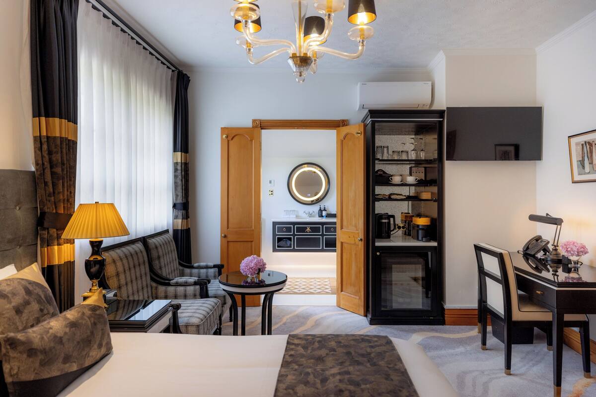 A hotel room with a bed, a sofa, a desk with a lamp, a chair, a dining table, and a kitchen area visible through an open door.