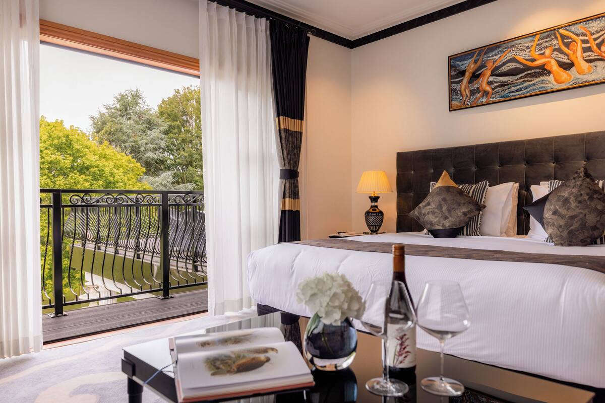 A hotel room with a large bed, a balcony with a view of trees, a painting on the wall, and a table with a vase and wine glass.