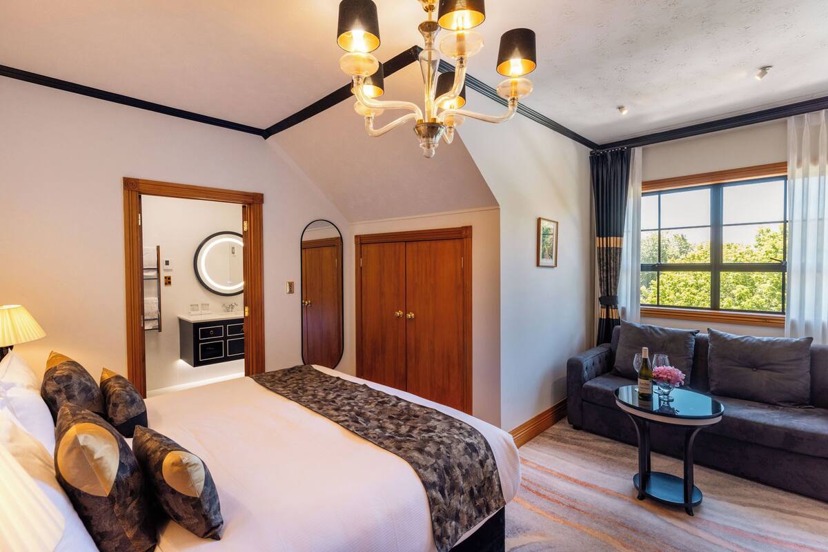 A hotel room with a large bed, a sofa, a small table with a bottle and glasses, a mirror, and a chandelier.