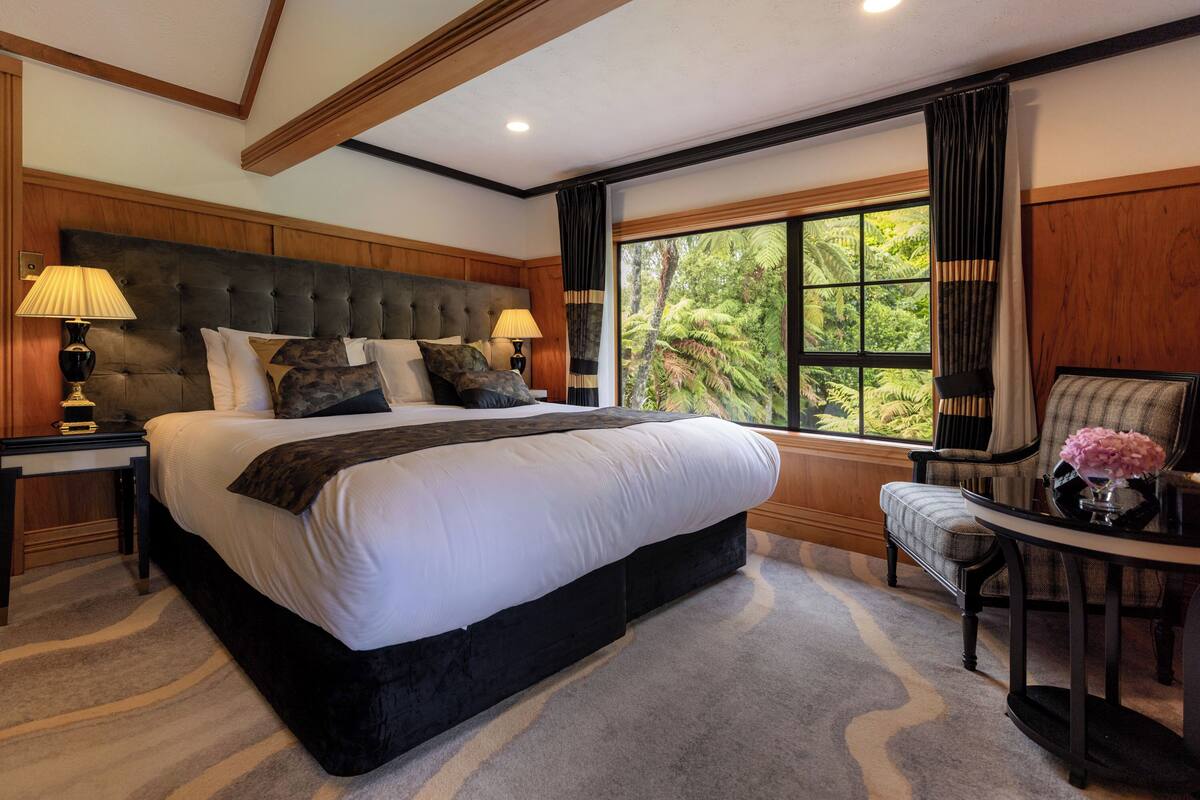 A large bed with a headboard, two bedside tables with lamps, a chair, and a window with a view of greenery.