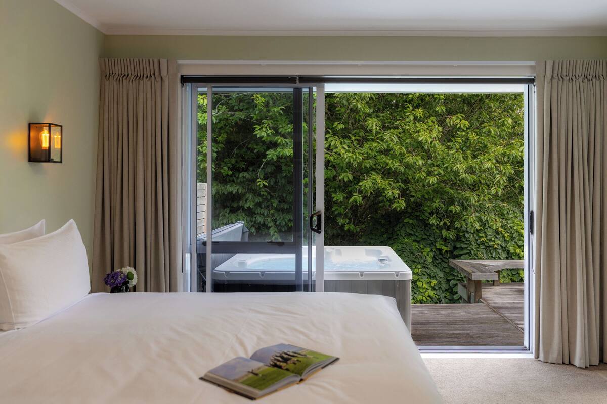 A hotel room with a large bed, a sliding glass door leading to a balcony with a hot tub, and a view of lush greenery.