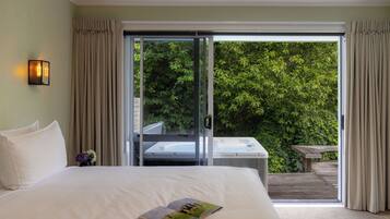 A hotel room with a large bed, a sliding glass door leading to a balcony with a hot tub, and a view of lush greenery.