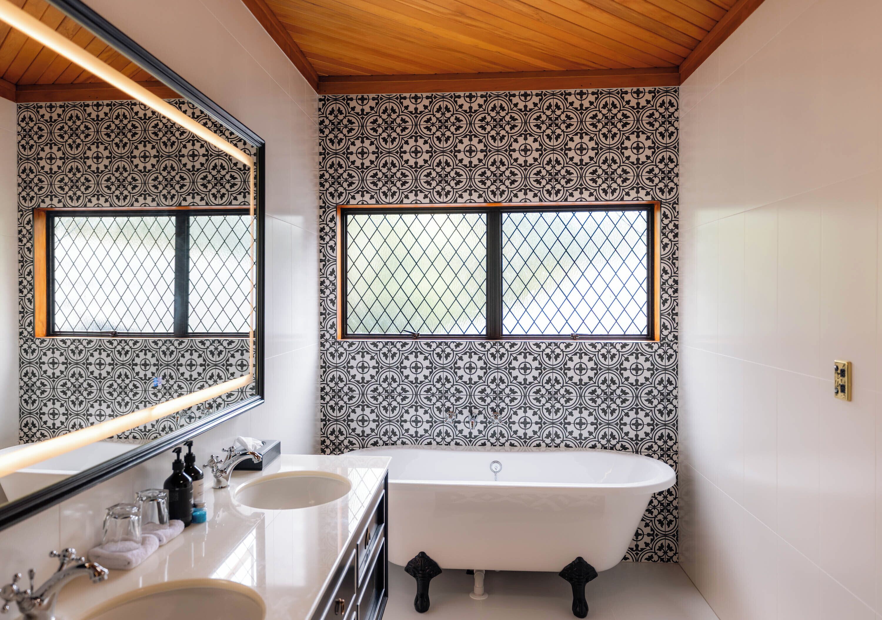 Un baño con una bañera independiente, dos lavamanos, un espejo grande y paredes con azulejos estampados.