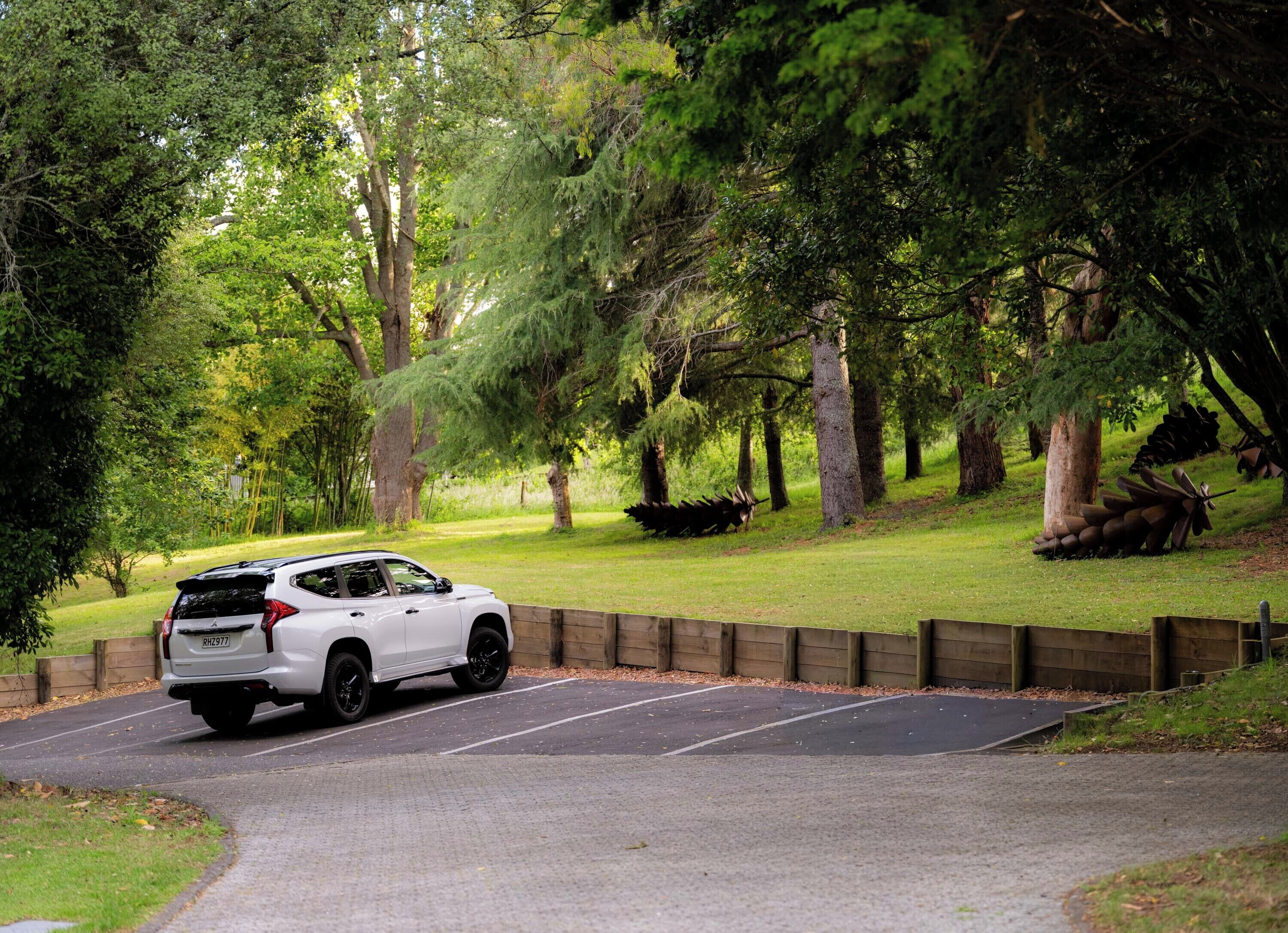 A white SUV parked on a paved path in a wooded area with a wooden fence and trees.