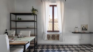 Un coin repas avec un sol à carreaux, une table dressée avec des assiettes et des verres, une bouteille de vin et une plante posée sur une étagère.