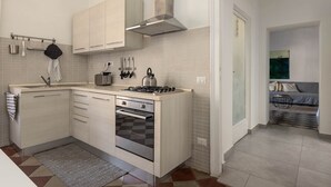 Une cuisine moderne dotée d’un four encastré, d’une cuisinière à gaz et d’une hotte en acier inoxydable. On y trouve un petit îlot central avec un évier et une bouilloire. La cuisine est agrémentée de meubles de couleur claire et d’un dosseret carrelé.