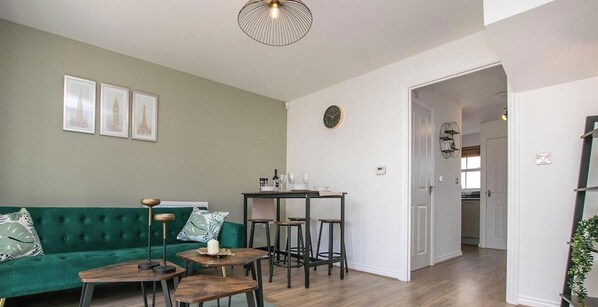 A living room with a green sofa, a dining table with chairs, a pendant light, and a clock on the wall.