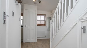 A hallway with white walls, a wooden floor, and a white door leading to another room. There is a window with blinds, a radiator, and a ceiling light fixture.