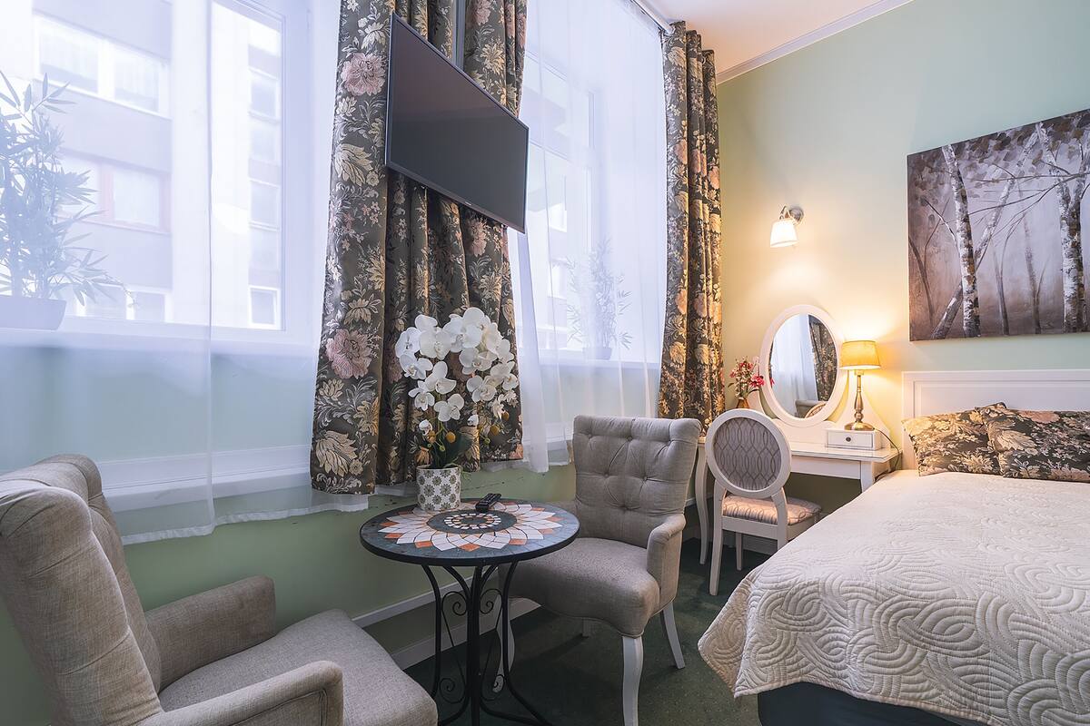 Une chambre d’hôtel comprenant un lit, deux fauteuils, une petite table avec un vase de fleurs, un téléviseur fixé au mur et un tableau accroché au mur.