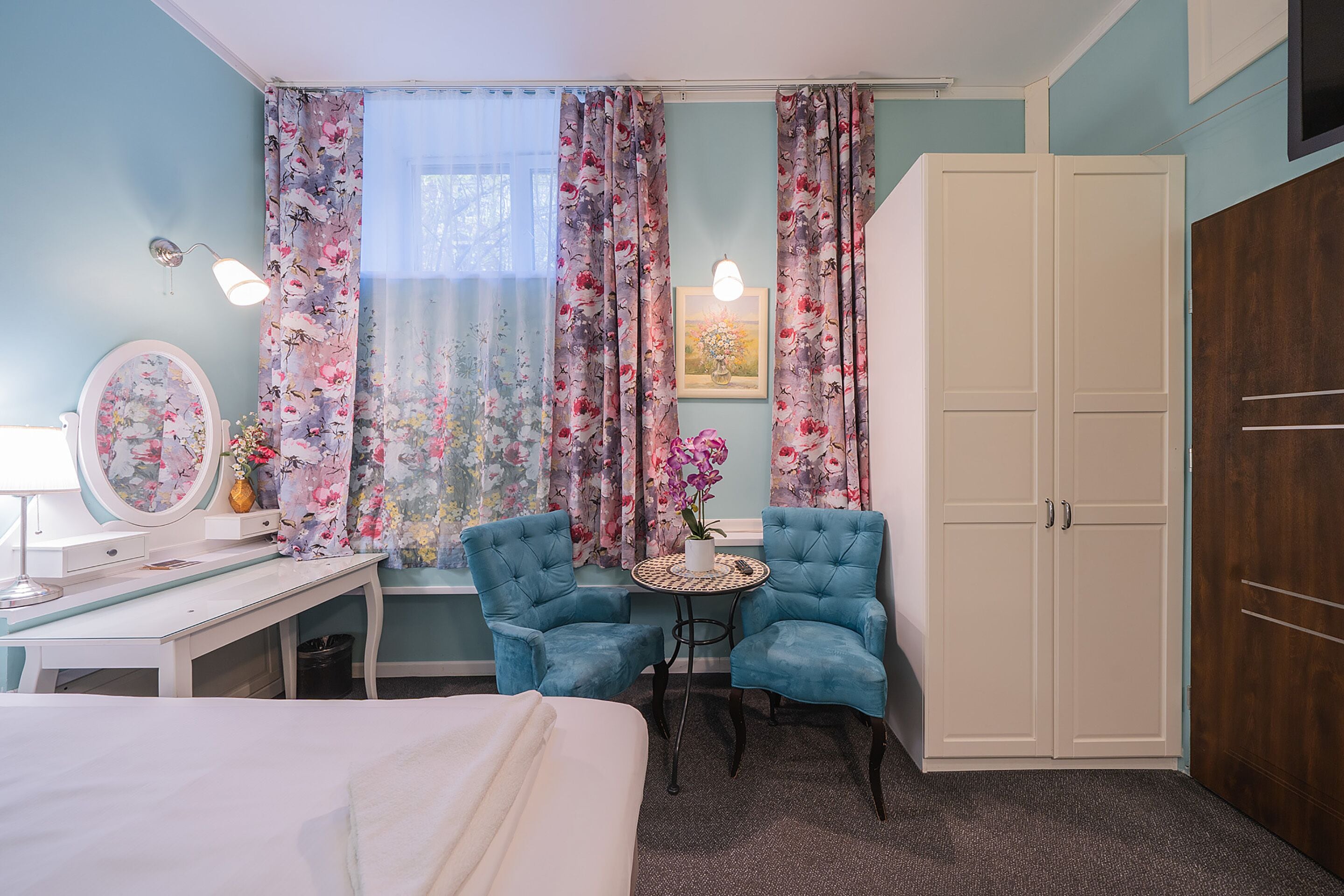 A bedroom with a bed, a desk, two chairs, a wardrobe, and floral curtains.
