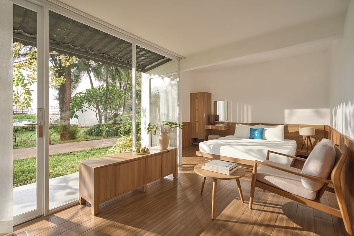 Un dormitorio moderno con un amplio ventanal corredero que da a un jardín, un banco de madera, una cama con almohada azul, una mesita de noche y una mesita auxiliar con lámpara.