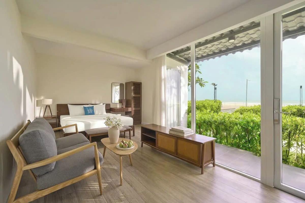 Habitación de hotel con cama, sofá, una mesita con un florero, armario y puerta corrediza de vidrio que da a un balcón con vistas a la vegetación y una playa.
