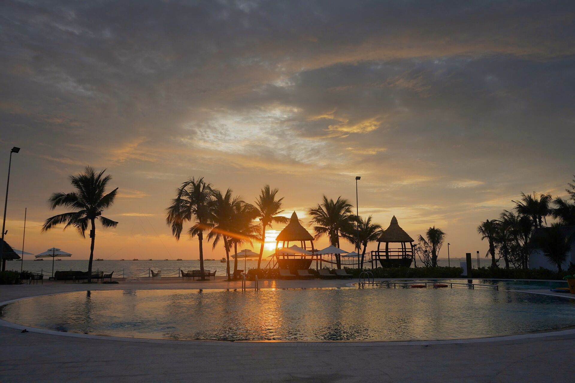 Área de piscina em resort com palmeiras e estruturas de telhado de palha ao pôr do sol.