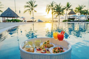 A poolside breakfast setup with a basket of food and drinks, palm trees, and a sunset in the background. - Palace Long Hai Resort (Ho Chi Minh City)