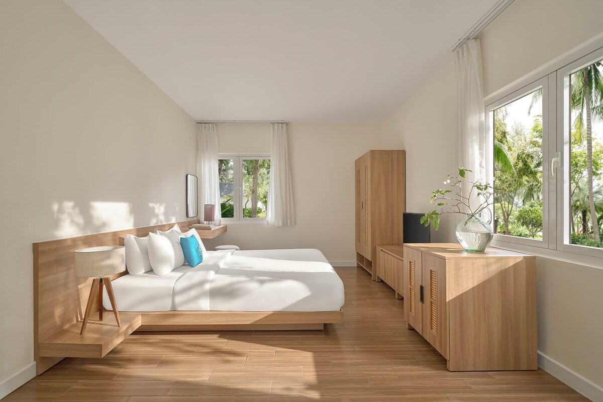 A modern bedroom with a large bed, wooden headboard, and a view of greenery through the window.