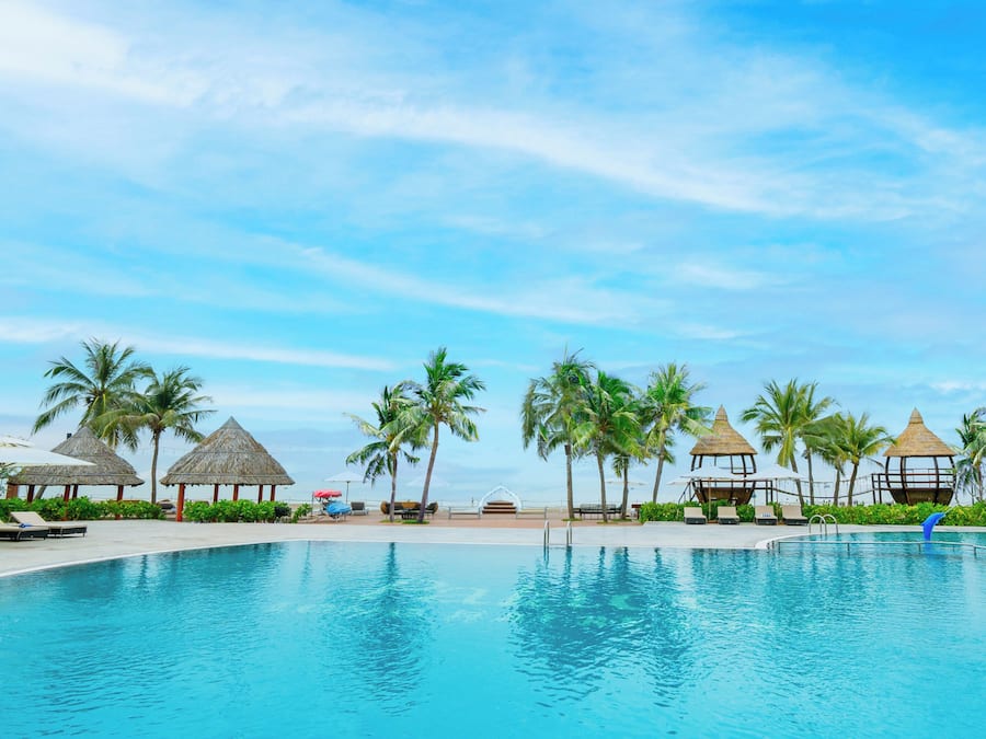 A swimming pool with lounge chairs and palm trees, surrounded by thatched-roof structures.