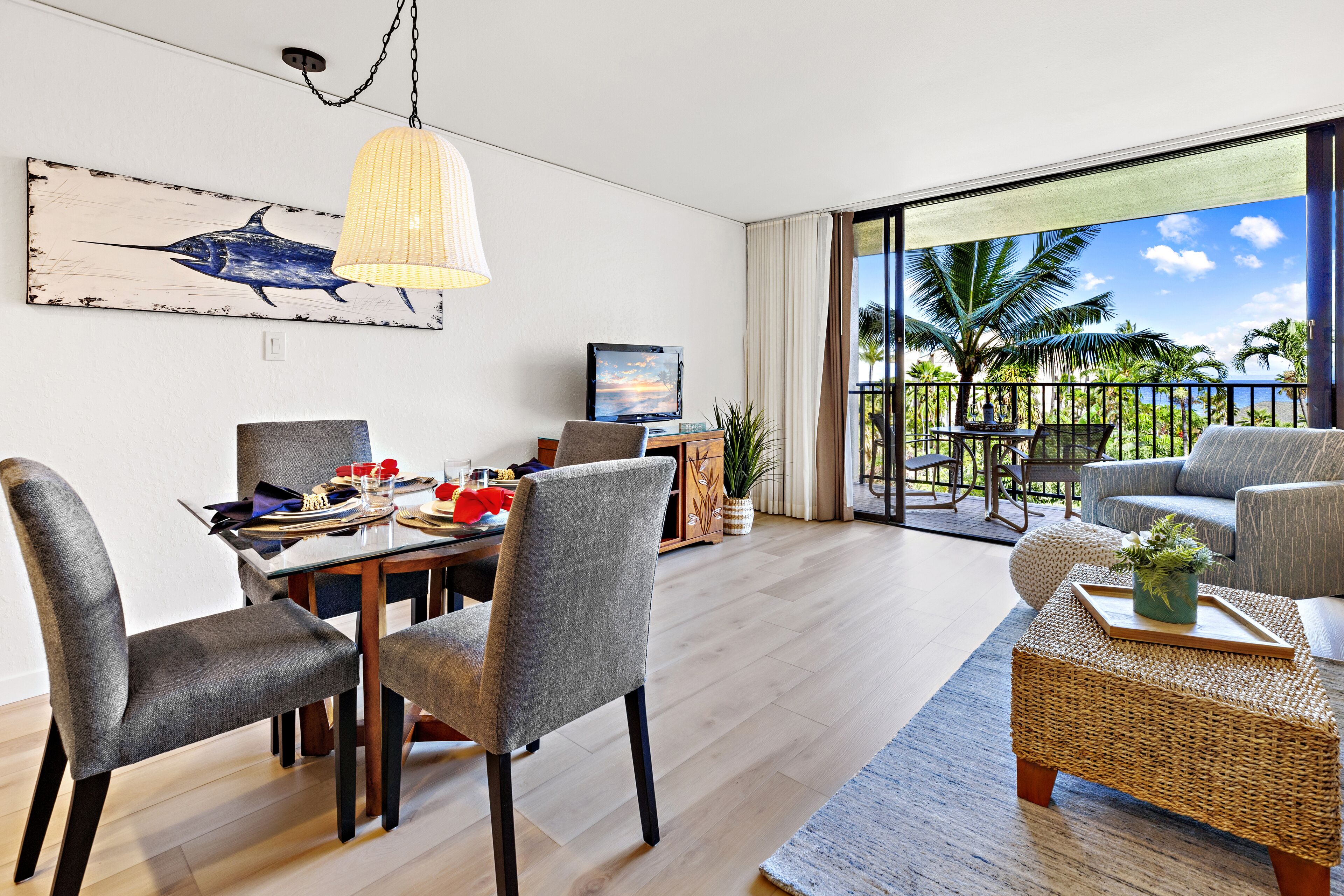A modern living room with a dining area, a large window with a view of palm trees and the ocean, and a mounted artwork of a blue fish.