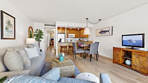 A modern living room with a sofa, coffee table, and TV stand. There is a kitchen in the background with wooden cabinets and a dining area. - Tropical charm, ocean touches - A guest favorite (Lahaina)
