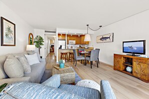 A modern living room with a sofa, coffee table, and TV stand. There is a kitchen in the background with wooden cabinets and a dining area.