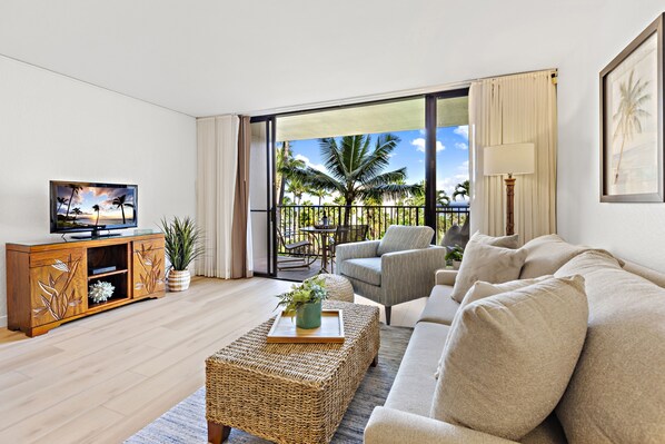 Un salon avec un téléviseur, un canapé et une table basse, un balcon avec vue sur les palmiers et un meuble en bois.