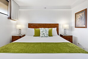 Une chambre d’hôtel bien rangée, avec un grand lit, deux tables de chevet avec des lampes, un plaid vert et une œuvre d’art encadrée accrochée au mur.