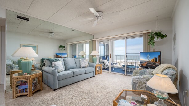 A spacious living room with a large grey sofa, a coffee table, and a television. There are two side tables with lamps, a ceiling fan, and a large window with a view of the city. - Direct oceanfront two bedroom condo ideally located on 77th street in Ocean City (Ocean City)