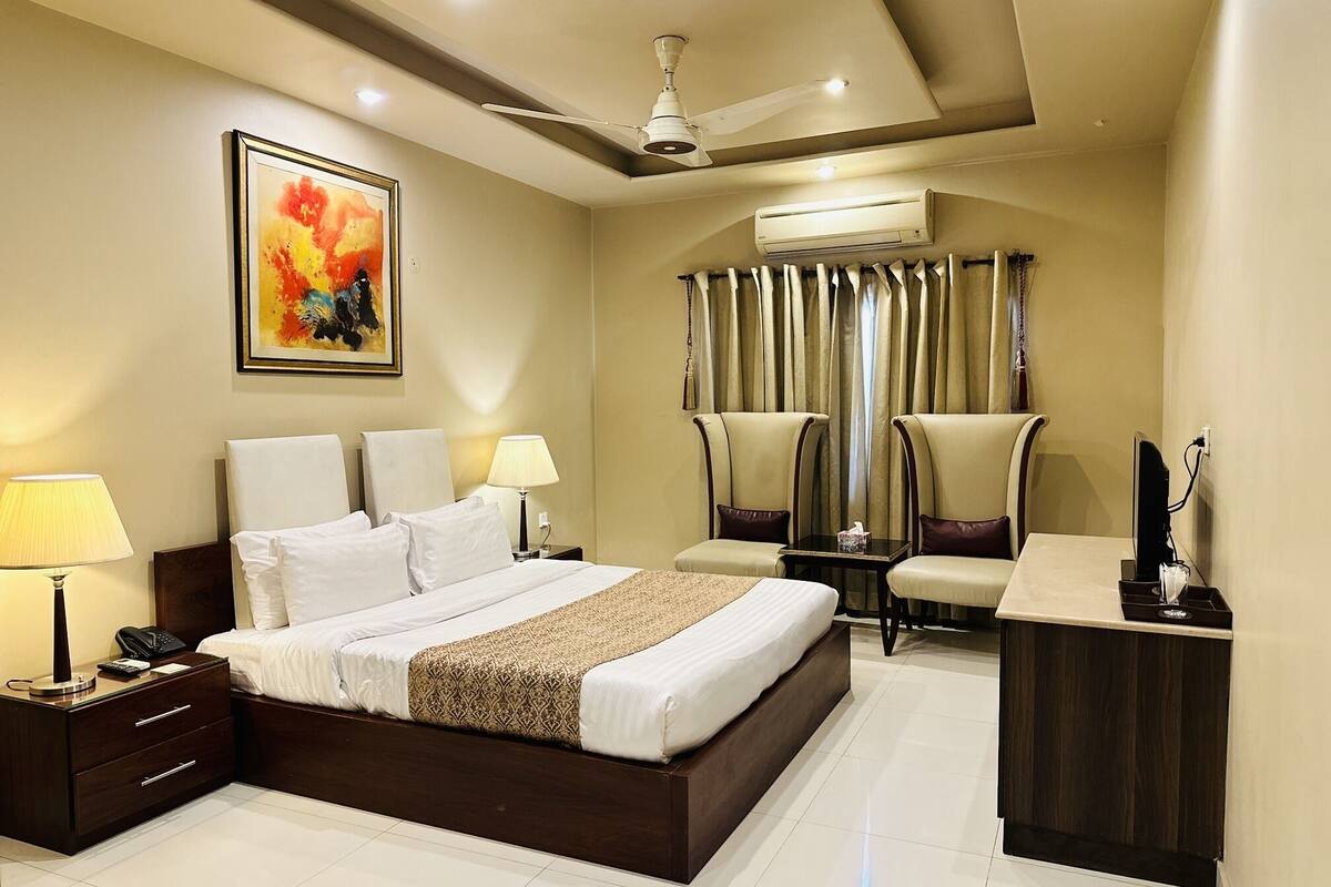 A hotel room with a large bed, two bedside tables, a desk, a chair, an air conditioning unit, and a ceiling fan.