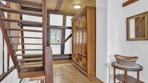 Een houten trap met een leuning, een grote houten garderobe, een ronde houten tafel met een mand, en een spiegel aan de muur.