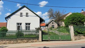 Une maison blanche à deux étages, avec une clôture verte, un mur de pierres et un portail en métal.