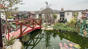 A serene garden with a red bridge over a pond, surrounded by various statues and plants.