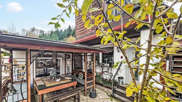 A covered patio area with a wooden table and chairs, a small seating area, and a view of a building with a red roof.