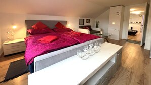 A modern bedroom with a large bed, red bedding, bedside tables with lamps, a glass coffee table, and a view into the kitchen.
