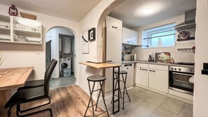 A compact kitchen with a washing machine, a dining area with a wooden table and chairs, and a small bar stool.