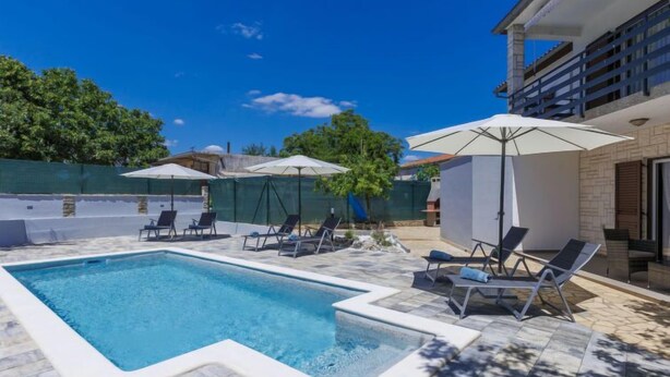 A swimming pool with lounge chairs and umbrellas, surrounded by a stone patio and a fenced area with trees. - Vacation home Reaa by Interhome (Barban)