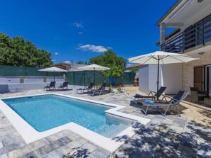 A swimming pool with lounge chairs and umbrellas, surrounded by a stone patio and a fenced area with trees. - Vacation home Reaa by Interhome (Barban)